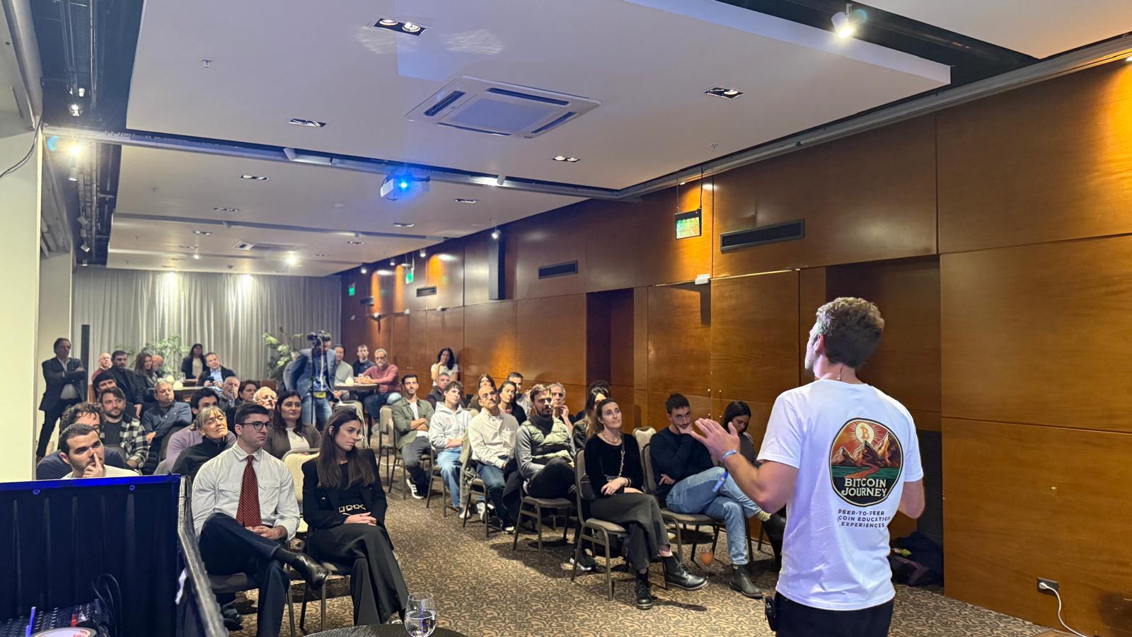 A speaker wearing a white Bitcoin Journey t-shirt presents to a seated audience inside a wood-panelled conference room